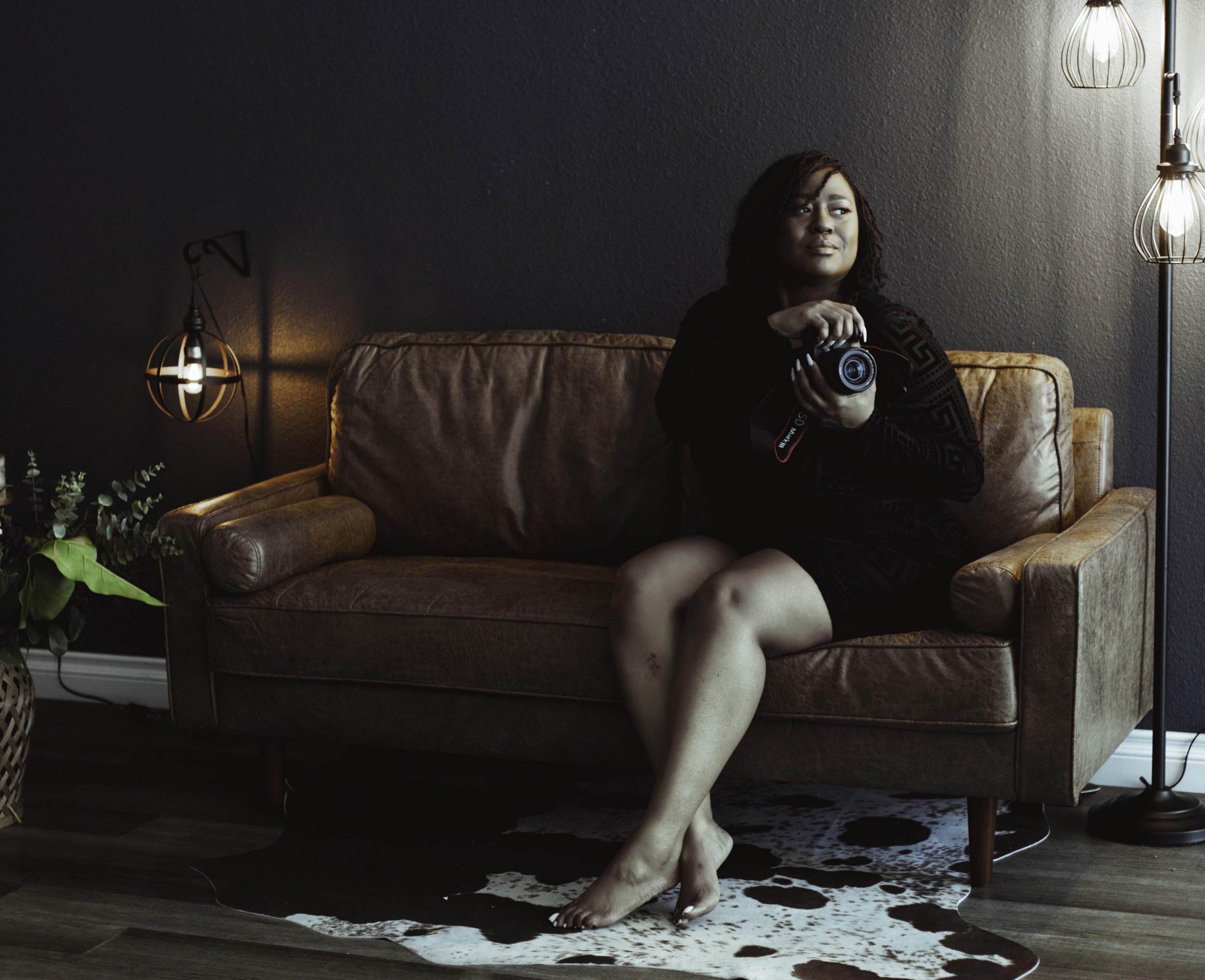 Latasha Nicole of Latasha Nicole Boudoir sits on a brown couch wearing a black dress holding a black Canon camera as she poses in her Vegas Boudoir Studio.