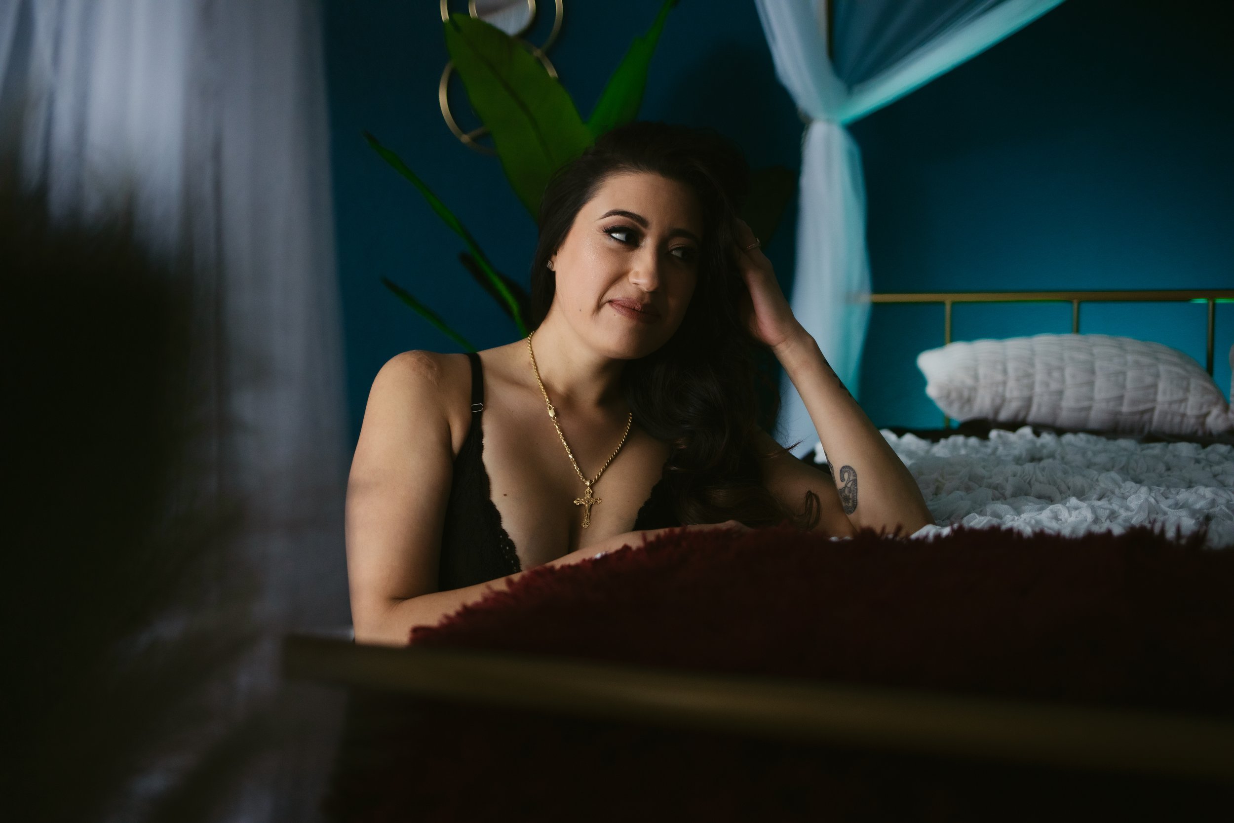 A woman with long dark hair sits on the side of a bed wearing a black gown, smiling coyly during a Las Vegas boudoir photoshoot